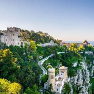 A stone castle sits atop a lush, green cliffside overlooking the ocean on a sunny day.