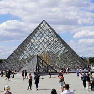 Una grande folla di persone passeggia attorno a una piramide di vetro in un ampio cortile pavimentato sotto un cielo parzialmente nuvoloso.