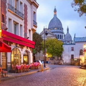 Una strada acciottolata deserta al crepuscolo, fiancheggiata da caffè ed edifici illuminati, con una grande basilica dalla cupola bianca sullo sfondo.