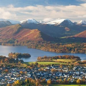 A high-angle view of a town on the shore of a large lake, with autumn-colored forests and snow-capped mountains behind it.