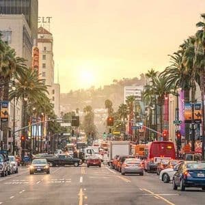 Un'ampia strada a Hollywood con traffico intenso, costeggiata da alte palme e edifici, sotto un tramonto dorato e velato.
