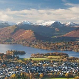 An aerial view of a lakeside town in autumn, with a backdrop of large, snow-capped mountains under a partly cloudy sky.