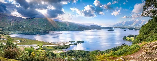 A panoramic view of a large lake surrounded by green mountains, with sunbeams breaking through dramatic clouds above.