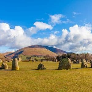 A circle of large standing stones sits in a grassy field with rolling hills and mountains in the background under a blue sky with white clouds.