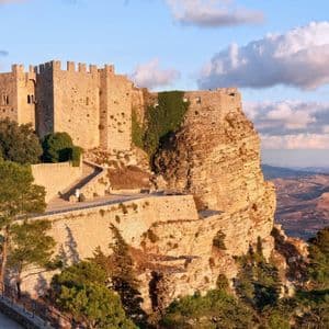 Un château de pierre perché sur une falaise rocheuse surplombant une vallée, illuminé par la lumière chaude du coucher de soleil.