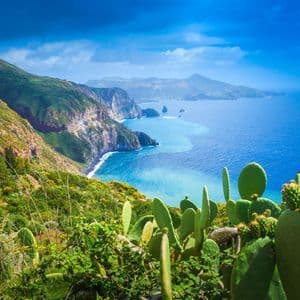 Vista dall'alto su fichi d'India, una costa verde e frastagliata, ripide scogliere e un mare turchese sotto un cielo blu drammatico.