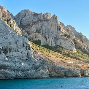 Une chaîne de montagnes accidentées et rocheuses, avec des pentes herbeuses, descend vers la mer calme et bleue sous un ciel clair.