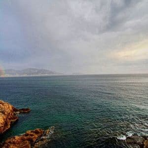 Un double arc-en-ciel enjambe la mer vers une ville lointaine, vu depuis une côte rocheuse sous un ciel nuageux.