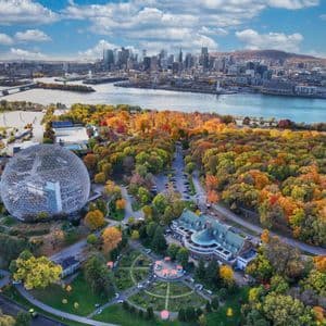 Vue aérienne d'un grand dôme géodésique dans un parc avec des arbres d'automne colorés, avec la silhouette d'une ville de l'autre côté d'une rivière en arrière-plan.