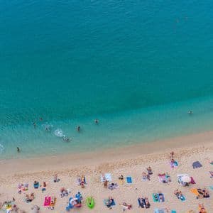 Una vista aerea di una spiaggia sabbiosa affollata con persone che nuotano in acqua turchese e prendono il sole su asciugamani colorati.
