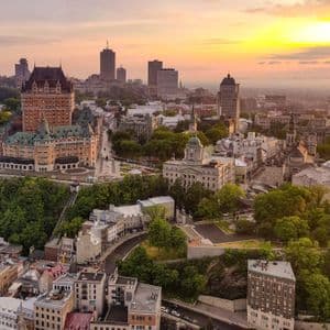 Vue aérienne d'une ville avec un grand hôtel aux allures de château et des bâtiments historiques nichés parmi les arbres, sous un coucher de soleil doré.