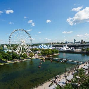 Une grande roue blanche et des tentes de cirque bordent un parc au bord de l'eau où des gens se promènent et font du bateau sous un ciel bleu.