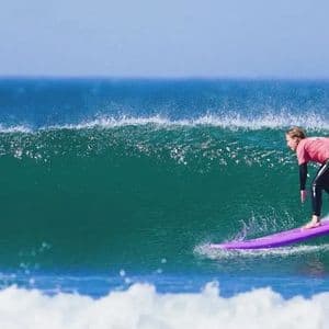 Un surfeur en chemise rose et combinaison chevauche une grande vague turquoise de l'océan sur un longboard violet sous un ciel bleu clair.