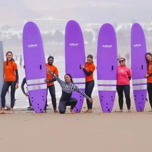 Un groupe WeRoad en voyage posant avec leurs planches de surf violettes et grises sur une plage de sable avant une leçon de surf.