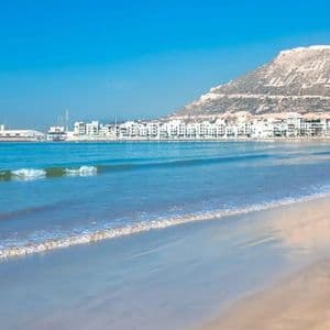Vue panoramique d'une vaste plage de sable fin où de douces vagues caressent le rivage, avec une ville côtière et une grande colline en arrière-plan sous un ciel bleu dégagé.