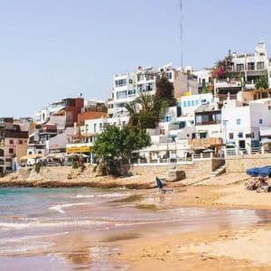 Une ville côtière aux bâtiments blanchis à la chaux est perchée sur une colline surplombant une plage de sable où les gens se détendent sous des parasols bleus.