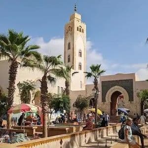 Une place de marché en plein air animée avec un haut minaret, des palmiers et des passants déambulant parmi les étals sous un ciel bleu et ensoleillé.