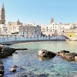 Vista soleggiata di una storica città costiera con edifici bianchi e un campanile, da una limpida baia turchese.