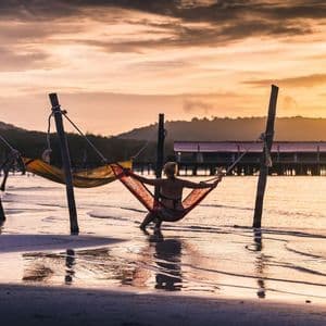 Une personne vue de dos est assise dans un hamac rouge au-dessus de l'eau sur une plage, lors d'un coucher de soleil doré.