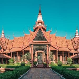 Un bâtiment orné, brun-rougeâtre, à l'architecture traditionnelle entoure une cour centrale avec des jardins manucurés et un chemin de pierre sous un ciel clair.