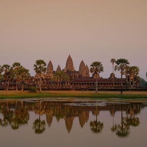 Un ancien complexe de temples avec des tours et des palmiers se reflète dans l'eau calme d'un lac au coucher du soleil.