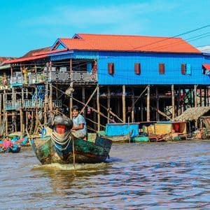 Un homme dans un bateau en bois navigue sur une rivière le long de maisons colorées sur pilotis dans un village flottant sous un ciel bleu clair.