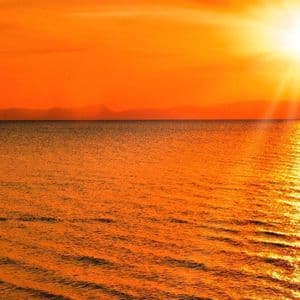 Une silhouette d'une personne debout dans la mer peu profonde au coucher de soleil orange éclatant, le soleil se reflétant sur l'eau.