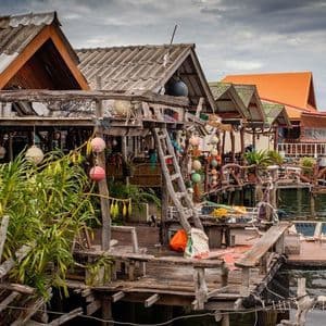 Un ensemble de maisons rustiques en bois sur pilotis construites sur l'eau, décorées de bouées colorées et de plantes sous un ciel nuageux.