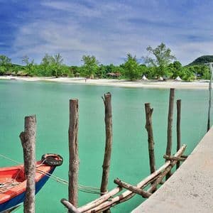 Une jetée en béton s'étire sur des eaux turquoises vers une plage de sable bordée d'arbres luxuriants, où un bateau rouge et bleu est amarré.