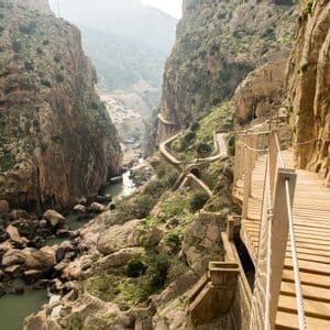 A narrow wooden boardwalk snakes along a sheer rock cliff face high above a river in a deep canyon.