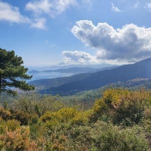Una vista panoramica da una collina che mostra verdi colline ondulate, un mare lontano e montagne sotto un cielo azzurro parzialmente nuvoloso.