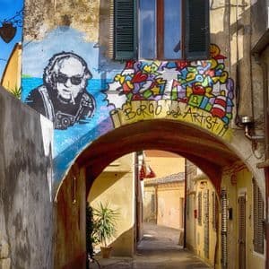 Graffiti colorati, incluse le parole 'Borgo degli Artisti', decorano un arco sopra uno stretto vicolo tra edifici gialli in una giornata di sole.