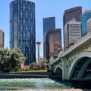 A modern city skyline with skyscrapers and an observation tower, viewed from across a river with a stone arched bridge.