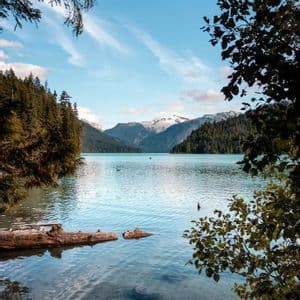 Un sereno lago alpino con agua cristalina, rodeado de densos bosques de pinos y montañas nevadas bajo un cielo parcialmente nublado.