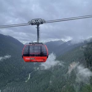 Una góndola roja Peak 2 Peak cuelga de cables muy por encima de un valle boscoso con montañas nevadas bajo un cielo nublado.