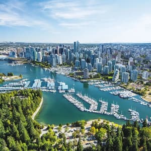 Una vista aérea de un horizonte urbano moderno junto a una bahía con marinas llenas de barcos y un denso bosque verde.