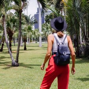 Eine von hinten gesehene Frau mit schwarzem Hut, roter Hose und Rucksack geht durch einen grasbewachsenen Park, umgeben von Palmen.