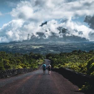 Zwei Männer mit Rucksäcken gehen eine Straße entlang durch einen üppigen grünen Wald auf einen großen, wolkenverhangenen Berg in der Ferne zu.