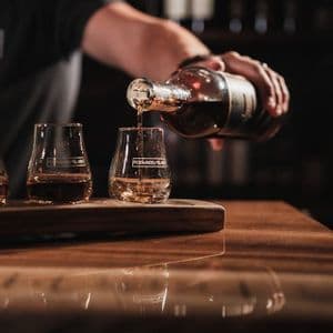 Eine Hand gießt Whiskey aus einer Flasche in ein Verkostungsglas, das auf einem Holztablett auf einer spiegelnden Bartheke steht.