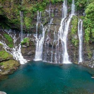 Mehrere Wasserfälle stürzen eine üppig grüne Felswand hinab in ein klares türkisfarbenes Becken, wo mehrere Leute schwimmen.