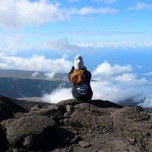 Eine langhaarige Person, von hinten gesehen, sitzt mit einer Mütze auf einem felsigen Gipfel und blickt über eine wolkengefüllte Schlucht.