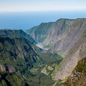 Luftaufnahme eines riesigen, grünen Canyons mit steilen Berghängen, einem Talboden darunter und dem Ozean am Horizont sichtbar.