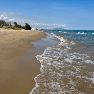 Wellen mit weißem Schaum rollen an einen Sandstrand unter einem klaren blauen Himmel, mit fernen Bergen am Horizont.