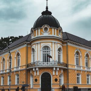 Un elaborato edificio angolare giallo con una cupola nera si erge su una strada di ciottoli bagnata sotto un cielo nuvoloso.
