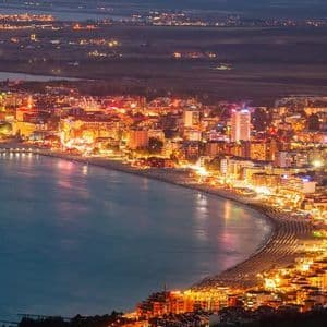 Vista aerea di una città costiera con edifici luminosi che fiancheggiano una spiaggia curva di notte.