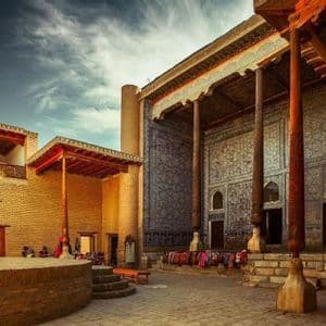 A sunlit courtyard with brick buildings, featuring large carved wooden pillars and a facade decorated with intricate blue geometric tiles.
