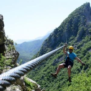 Ein Mann mit gelbem Helm fährt mit der Seilrutsche über ein tiefes Tal zwischen zwei grünen, felsigen Bergen unter klarem Himmel.