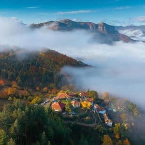 Vue aérienne d'un petit village à flanc de montagne, couvert d'arbres aux couleurs automnales, émergeant d'une mer de nuages.