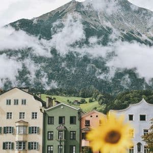 Eine Reihe bunter Gebäude steht am Fuße eines großen, wolkenverhangenen Berges, mit einer gelben, unscharfen Sonnenblume im Vordergrund.