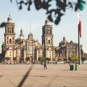 Une grande cathédrale historique avec des clochers jumeaux se dresse sur une place ensoleillée où les gens se promènent, avec un drapeau mexicain flottant à droite.